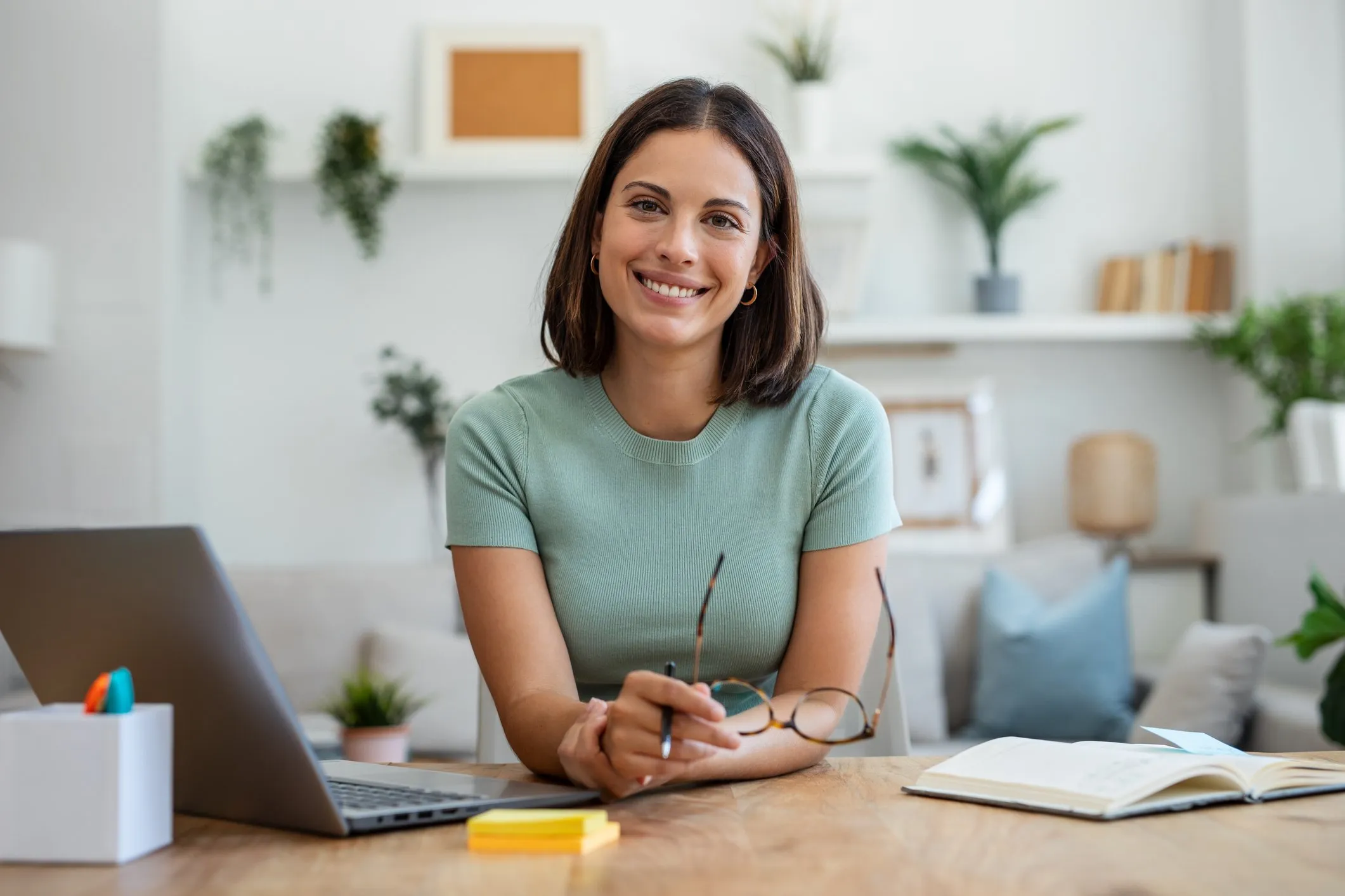 A woman with shoulder-length brown hair, wearing a light green t-shirt, smiling and holding glasses, sitting at a wooden desk with an open book, a closed laptop, and some sticky notes in a well-lit, cozy room with plants and shelves in the background.