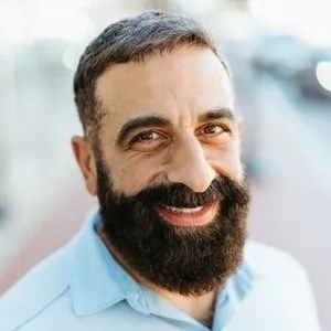 Close-up of a man with dark hair and a full beard, smiling, wearing a light blue shirt with a blurred outdoor background.