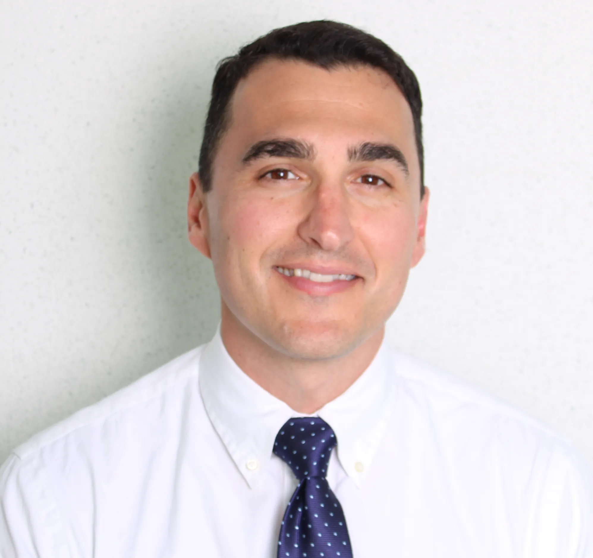 Portrait of a young man with dark hair, dressed in a white shirt and navy blue polka dot tie, smiling against a plain light background.