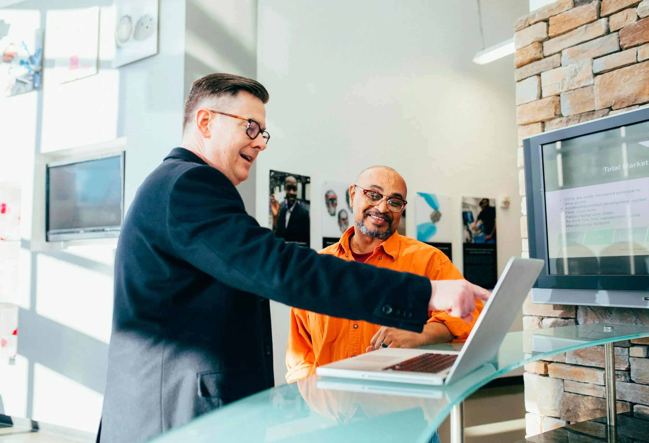 Two men, one wearing a black blazer and glasses and the other in an orange shirt, are standing at a glass reception desk. They are smiling and looking at a laptop, with the man in the blazer pointing at the screen. There are computers and artwork on the walls in the background.